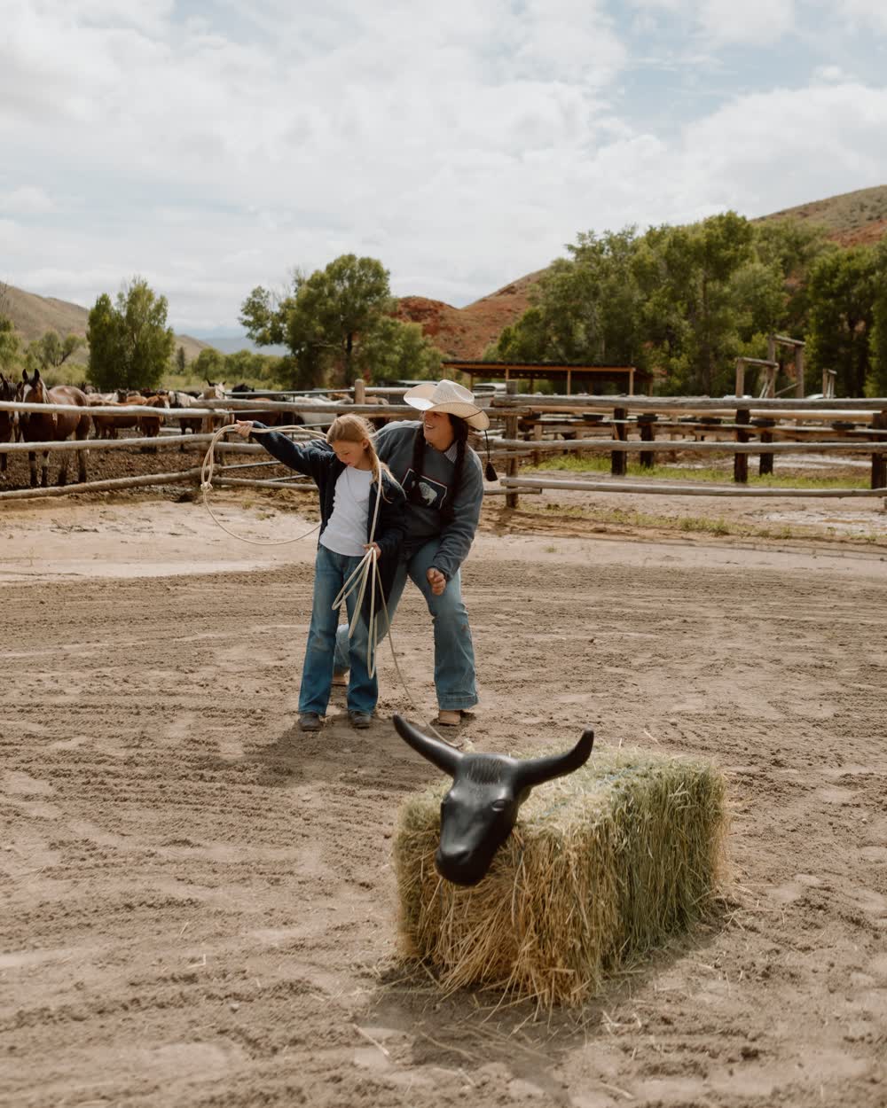 A child learning to rope at a ranch corral, horses waiting behind the rail
